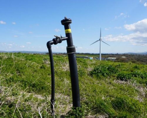 Landfill gas pipe sticking out of the ground with a wind turbine in the background
