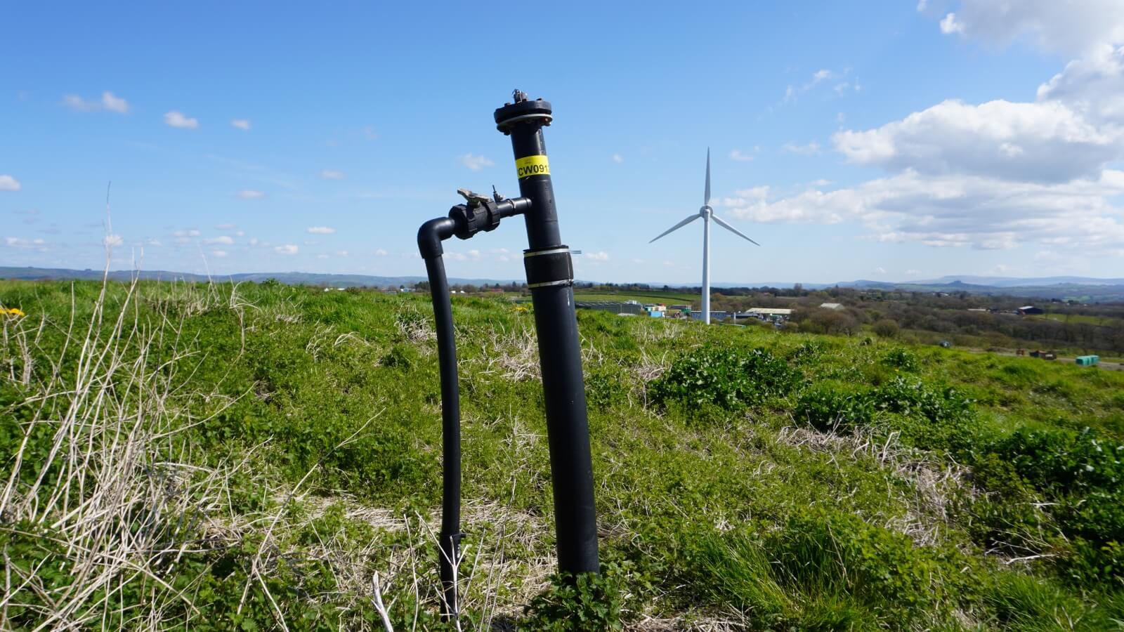 Landfill gas pipe sticking out of the ground with a wind turbine in the background