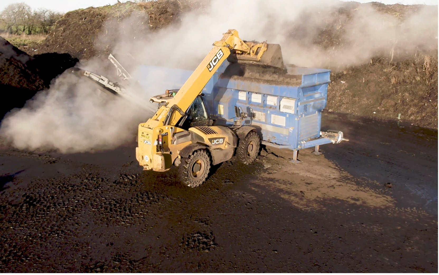 A JCB grabber dropping green waste into an industrial device